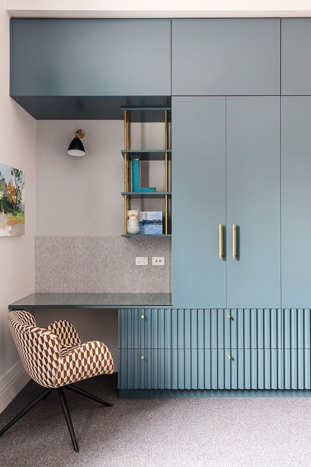 A compact, carefully designed home office with extensive muted blue-grey built-in cabinetry, vertically fluted drawer fronts, brass handles, and a dark stone or composite desktop. An open shelving niche displays books and objects beside a wall-mounted black-and-brass sconce. A patterned upholstered desk chair sits on a grey carpet, with soft neutral walls and a small artwork adding colour.