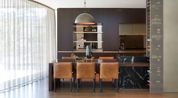 Open-plan dining and kitchen interior with a long timber dining table and three leather-upholstered chairs in the foreground. A dark wood kitchen with integrated cabinetry, open shelving, dark stone benchtops and a dramatic black marble island forms the backdrop. A large dome pendant hangs above the table, while sheer full-height curtains provide soft daylight along the glazed wall.