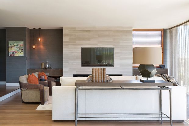 A bright contemporary living room viewed from behind a large white sectional sofa. The room features a pale horizontal timber-look television feature wall, dark charcoal built-in cabinetry and wall panelling, warm timber flooring, expansive sheer-curtained glazing, and layered neutral furnishings. A sculptural dark lamp and orange cushions provide accents, while a slim metal console sits behind the sofa.