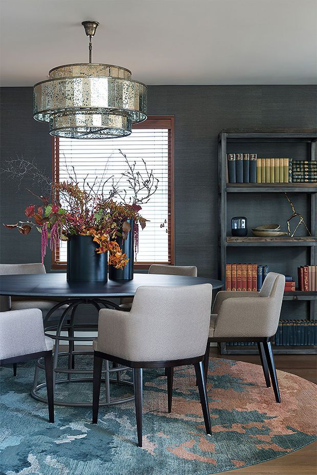 A refined dining room with a round dark dining table, upholstered dining chairs, a large patterned area rug, and a dramatic tiered metallic pendant light. Dark textured walls, a timber-framed window with horizontal blinds, a full-height open bookshelf filled with books, and sculptural floral arrangements create a moody, layered interior.