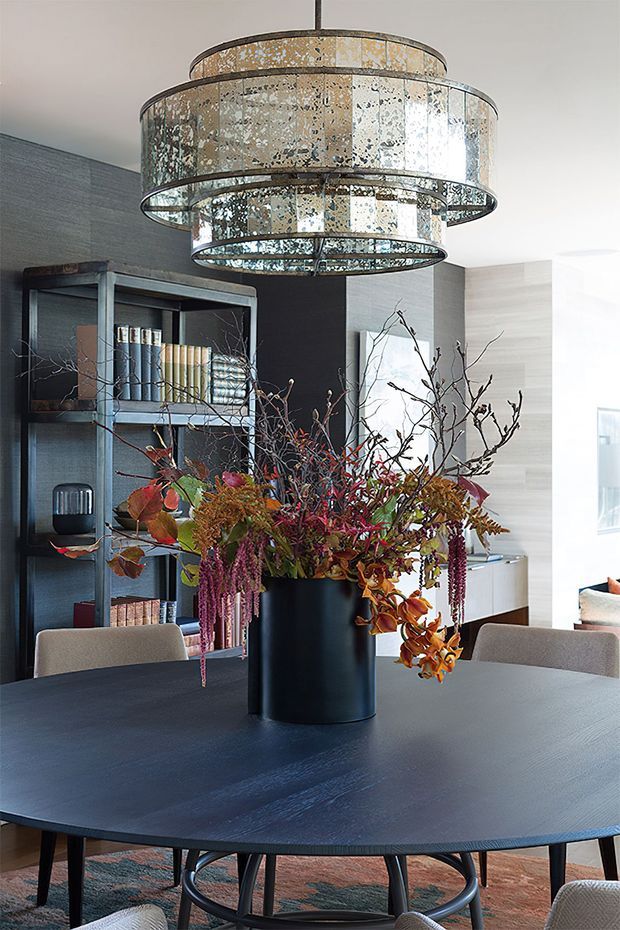 A sophisticated dining area with a large dark dining table, upholstered dining chairs, a black metal shelving unit, and a dramatic multi-tiered mirrored glass pendant light. A tall black vase filled with an abundant arrangement of dried and autumnal branches forms the table centrepiece. The room combines dark textured walls, light timber-toned cabinetry, and refined contemporary furnishings.