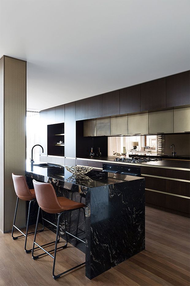 A sophisticated contemporary kitchen with a large black veined stone island and integrated breakfast bar seating. Dark timber cabinetry runs along the rear wall, paired with metallic upper cabinet fronts, a reflective backsplash, black fixtures, and warm timber flooring. The space is open, refined, and strongly contrasted, with a tall textured partition at the side and natural light entering from the rear.