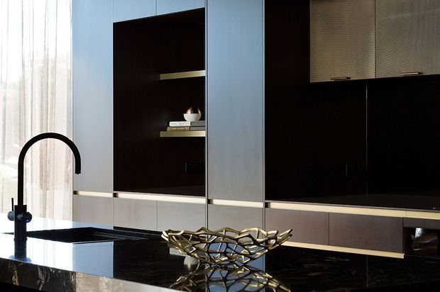 A refined contemporary kitchen with a large glossy dark stone island, integrated black gooseneck tap and sink, pale wood-toned lower cabinetry, and tall dark built-in cabinetry with open display shelving. A sculptural metallic bowl sits on the island, while sheer curtains provide soft daylight at the window.