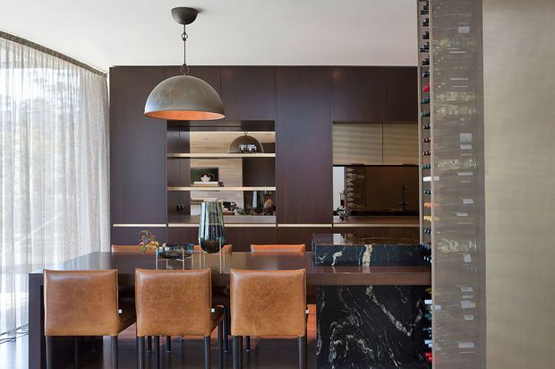 A sophisticated open-plan dining and bar area with a dark timber built-in wall, integrated shelving, marble-faced cabinetry, and a long dining table with brown leather upholstered chairs. A large dome pendant light hangs above the table, while sheer curtains provide soft natural light at the window. A tall glass-fronted wine display is visible at the right.