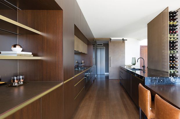A refined contemporary kitchen with dark timber cabinetry and integrated open shelving, brass-toned trim details, a long central island with a dark stone benchtop, and warm timber flooring. The space includes a dining overhang with leather upholstered stools, a sculptural pendant light, a wine display wall, and a compact galley-style layout extending toward a rear hallway.