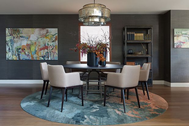 A refined dining room with a large round dining table surrounded by upholstered dining chairs, set over a circular blue and rust-toned area rug. Dark textured wall panelling, timber flooring, built-in open shelving, colourful abstract artwork, a central window, sculptural floral arrangements, and a large decorative pendant light create a sophisticated, layered interior.