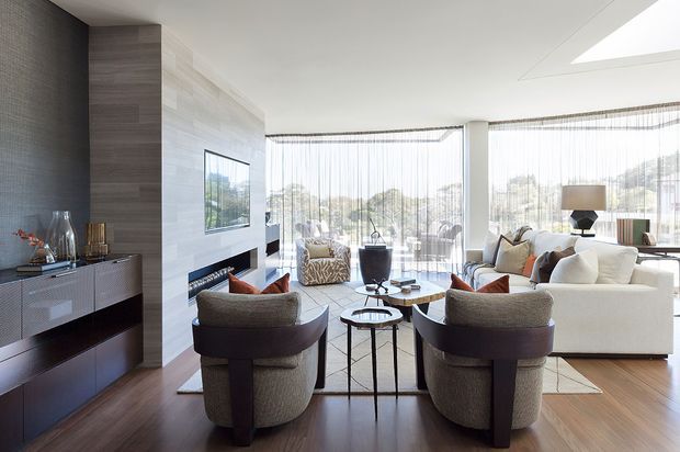 Bright, sophisticated contemporary living room with floor-to-ceiling sheer-glazed windows overlooking trees and hills. The room features dark timber flooring, a full-height textured feature wall and fireplace surround, built-in dark cabinetry, a large cream sofa, sculptural lounge chairs, glass coffee tables, and a skylight. The layout is open, airy, and refined.