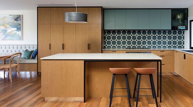 A contemporary open-plan kitchen with extensive warm timber cabinetry, a large central island with integrated seating, and a distinctive geometric circular-pattern tiled splashback. Muted blue-green upper cabinets contrast with the timber joinery, while a dining area and built-in upholstered bench are visible at left. Timber flooring, a sculptural pendant light, and clean-lined detailing create a refined, warm interior.