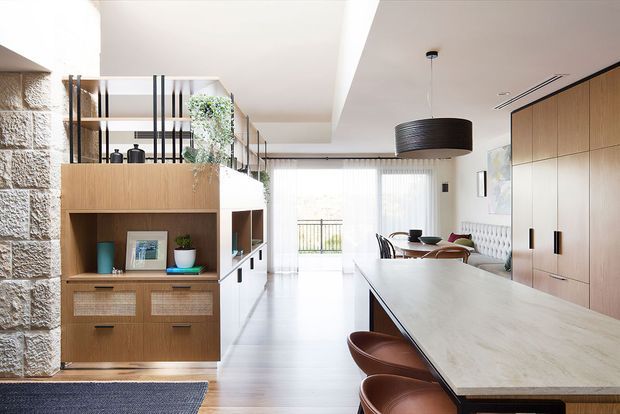 Bright open-plan contemporary interior with a kitchen island in the foreground, integrated timber cabinetry along the right wall, and a dining and living area beyond. A large pale stone island benchtop with brown bar stools dominates the space. Built-in timber shelving, black metal vertical detailing, textured stone wall cladding, pendant lighting, and full-height glazing create a warm, refined modern scheme.