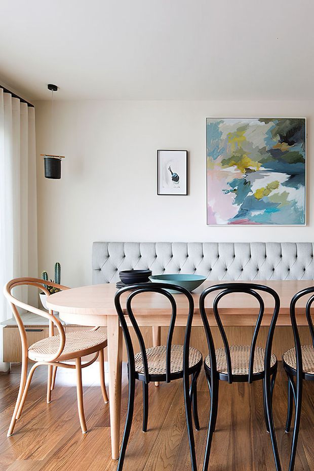 Bright dining area with a long light-wood dining table, black bentwood chairs, and a built-in tufted banquette. The room has warm timber flooring, white walls and ceiling, full-height curtains, potted plants, and colourful abstract artwork.