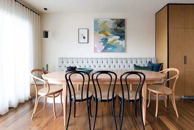 Bright, refined dining area with a large oval timber dining table surrounded by black bentwood-style chairs and woven-seat armchairs. A tufted pale upholstered banquette runs along the rear wall beneath abstract artwork, with tall sheer curtains on the left and full-height timber cabinetry on the right. Warm timber flooring and soft neutral finishes create a polished mid-century-inspired interior.