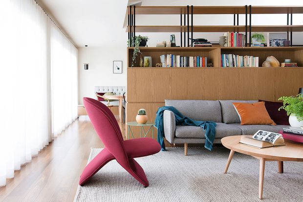 Bright, refined living room with floor-to-ceiling sheer curtains, pale timber flooring, a grey sofa, a bold magenta sculptural lounge chair, and a round timber coffee table. A full-width built-in timber shelving and cabinetry wall provides storage and display, with black vertical metal supports and colourful books and plants. A compact dining area is visible in the background.