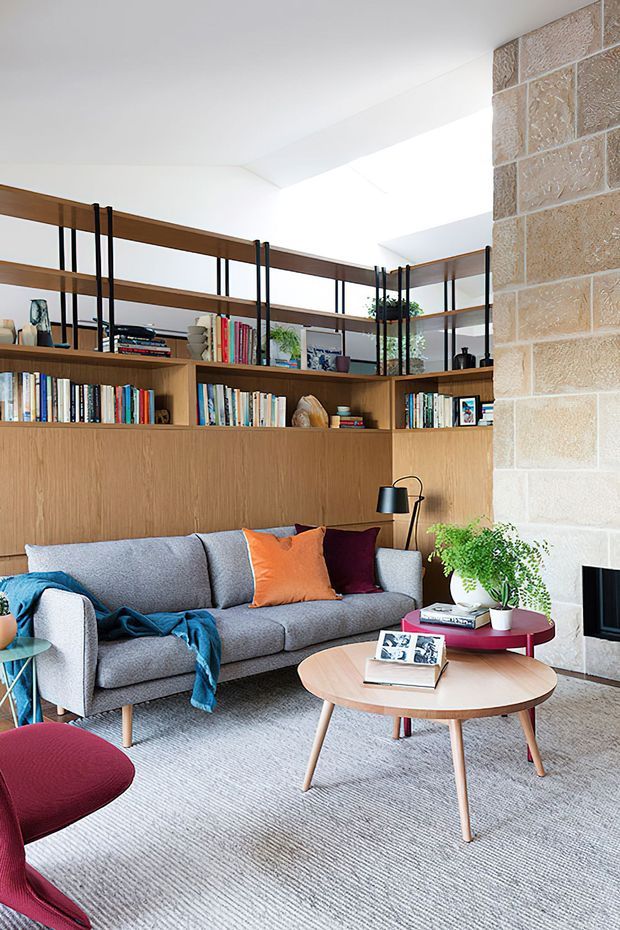 A bright, thoughtfully designed living room with a grey upholstered sofa, colourful cushions, a round timber coffee table, and a red accent chair. A large built-in timber bookcase with black metal supports spans the rear wall, while a textured pale stone feature wall and fireplace frame the right side. The room has a light carpet, abundant books and accessories, indoor plants, and a clean contemporary mid-century-inspired feel.