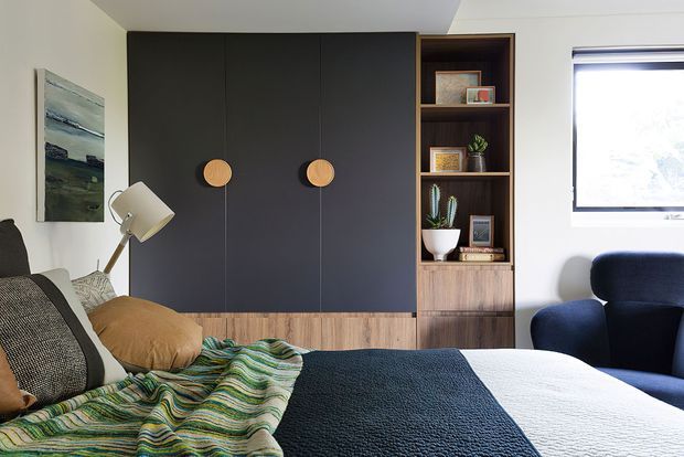 A warmly styled bedroom with a bed in the foreground, dark built-in wardrobe doors with round timber handles, and an integrated timber open shelving unit displaying books and decor. A navy armchair, floor lamp, framed artwork, and window complete the contemporary interior.