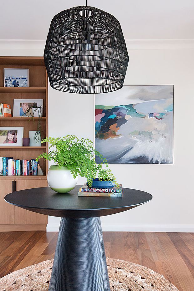 A bright, styled interior vignette featuring a round black pedestal dining table with a textured conical base, set on a woven rug. A large black woven pendant light hangs above, while a timber built-in bookshelf displays books, framed photos and decor. A colorful abstract artwork, potted greenery and timber flooring complete the space.