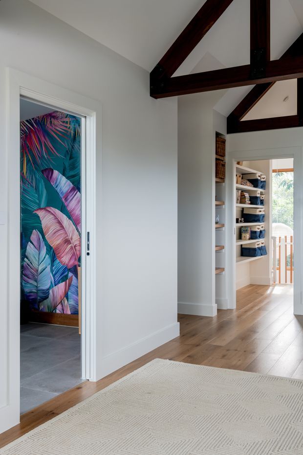 A bright residential hallway or landing with white walls, warm timber flooring, exposed dark timber roof trusses, and built-in open shelving leading toward a glazed exterior door. A colorful botanical feature wall is visible through a side doorway, while a large neutral patterned rug occupies the foreground.