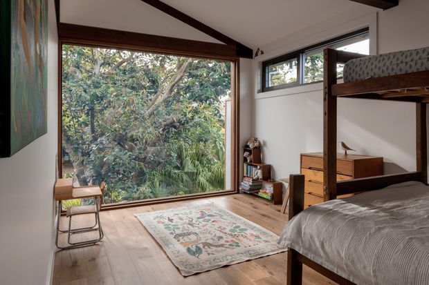A compact, light-filled bedroom with a timber bunk bed, wood flooring, a large timber-framed picture window overlooking dense greenery, and high horizontal clerestory-style windows. The room includes a small timber dresser, open shelving with books, a chair, patterned area rug, and exposed dark timber ceiling beams. The styling is warm, simple, and natural.
