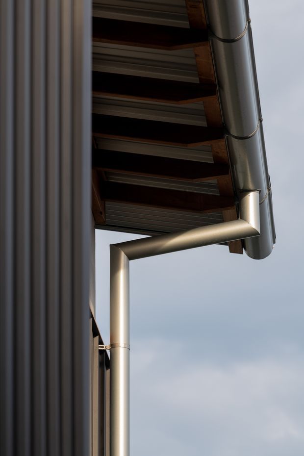 Close-up exterior architectural detail showing a metal downpipe and gutter connected beneath a roof eave. The eave reveals dark timber rafters with corrugated metal roofing or lining, alongside a dark vertical wall cladding. The image is tightly framed against an overcast sky and focuses on the junction and drainage hardware.