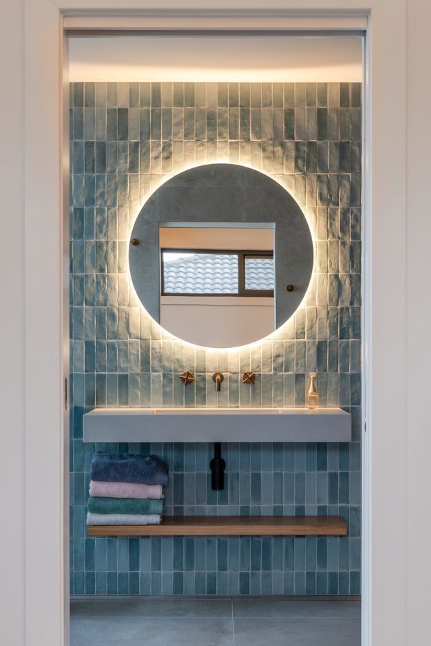 A compact bathroom vanity viewed through a doorway, featuring blue-green vertically arranged wall tiles, a floating white basin with exposed black plumbing, a warm timber shelf, folded towels, and a large round backlit mirror. Brass wall-mounted tapware and soft ambient lighting create a refined contemporary look.