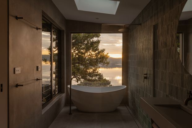 Moody contemporary bathroom featuring a freestanding sculptural bathtub positioned before floor-to-ceiling glazing, with a serene waterfront sunset view. The room has dark textured wall finishes, a skylight, minimalist black fixtures, a wall-mounted basin, and a frameless glass shower enclosure.