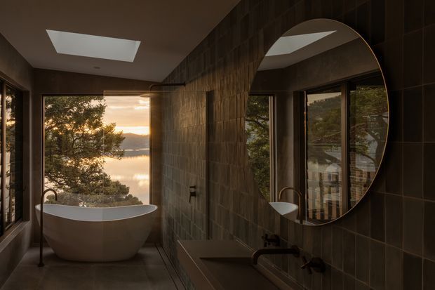 A moody, refined bathroom with a freestanding white bathtub positioned beside large windows overlooking a calm waterside landscape. Dark textured wall tiles, a broad stone or concrete vanity, black fixtures, a large rounded mirror, and a skylight create a dramatic, warm atmosphere. Timber-framed glazing and natural views add an organic connection to the exterior.