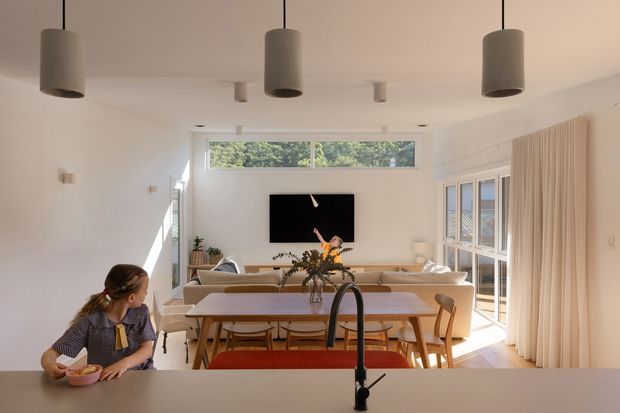Bright, open-plan contemporary living and dining area viewed from a kitchen island. The room features white walls and ceiling, expansive glazed doors and windows with full-height curtains, a neutral sectional sofa, timber dining table and chairs, and a black wall-mounted television. A dark kitchen tap and island bench occupy the foreground, while a child sits at the island. Simple cylindrical pendant lights and abundant natural light create an airy, warm interior.