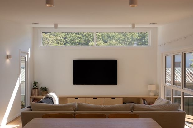 Bright, contemporary open-plan living and dining area with a wall-mounted television, low timber media cabinetry, large upholstered sofa, dining table, and extensive glazing. High horizontal windows and side windows provide abundant natural light and views of greenery and neighbouring rooflines. The room has a warm neutral palette and clean-lined built-in joinery.