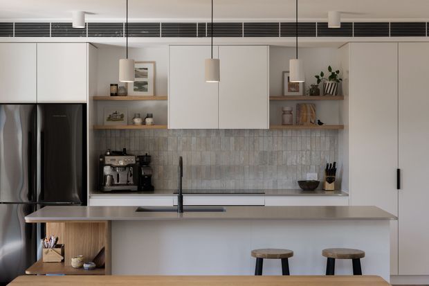A contemporary galley-style kitchen viewed from the dining area, featuring a large island with a pale stone benchtop, white flat-panel cabinetry, integrated black appliances, open timber shelving, and a vertically textured grey tile splashback. Three pendant lights hang above the island, with timber stools in the foreground and a long linear air-conditioning grille near the ceiling.