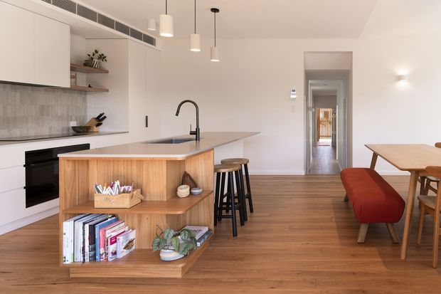 Bright open-plan kitchen and dining area with white flat-panel cabinetry, a pale tiled splashback, timber flooring, and a timber-lined island featuring open shelving. The island includes a dark benchtop, undermount sink, and curved black tap, with stools along the kitchen side. A timber dining table with upholstered rust-red seating occupies the adjoining space, while a hallway extends beyond.