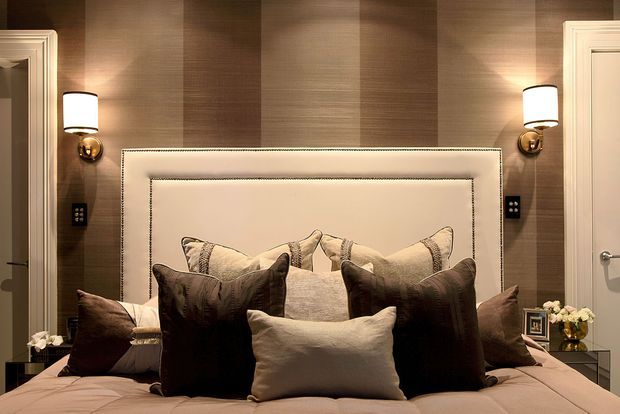 A luxurious bedroom viewed straight on, centered on a large upholstered bed with layered neutral and dark brown cushions. The room features warm vertical wood wall panelling, a padded cream headboard with nailhead trim, matching wall sconces, framed doorways on both sides, and subdued ambient lighting.