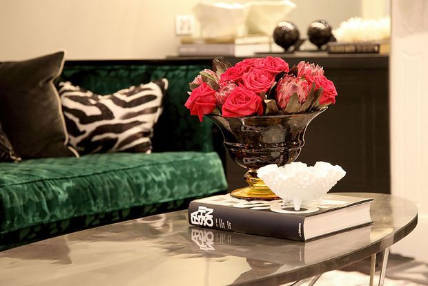 A close-up interior vignette showing a deep green velvet sofa with contrasting black-and-white cushions, a reflective oval coffee table, and a dramatic floral arrangement in a dark glass bowl placed on stacked books. Warm ambient lighting and glossy surfaces create a sophisticated, richly styled atmosphere.