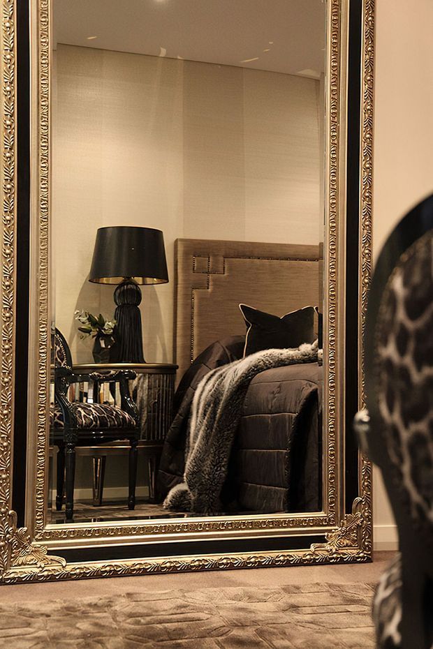 A warm, moody bedroom seen primarily through a large ornate gilt-framed floor mirror. The reflection shows a dark upholstered bed with a paneled headboard, black bedside lamp, small side table, patterned chair, and layered dark bedding with a plush throw. Neutral textured walls and a soft patterned carpet reinforce the luxurious, dramatic atmosphere.