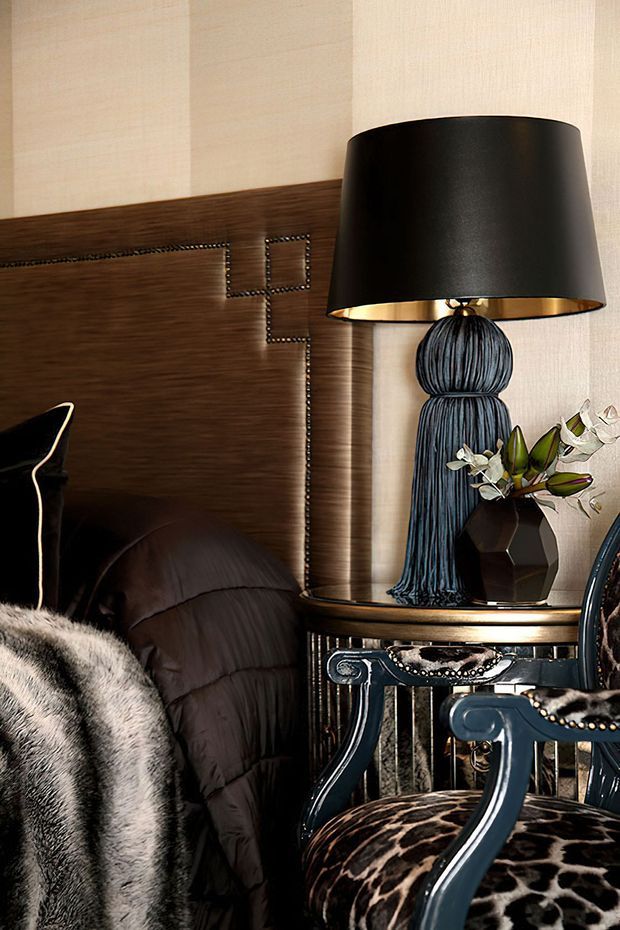 A richly styled bedroom corner featuring a dark wood, upholstered headboard with decorative nailhead trim, a black table lamp with a sculptural blue-grey base, and a round side table with metallic detailing. A dark upholstered chair with animal-print upholstery and a plush faux-fur throw adds dramatic texture. The palette is dark, warm, and luxurious with layered patterns and refined decorative accents.