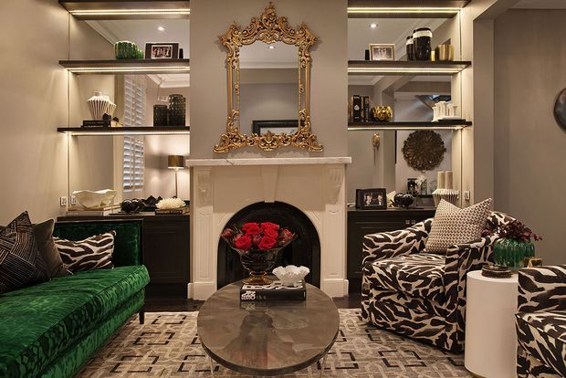 A richly styled living room centred on an ornate white fireplace and elaborate gold-framed mirror. Built-in dark shelving with integrated lighting displays decorative objects and framed photographs, while a deep green sofa and patterned cream-and-black armchairs surround a round glass coffee table. The room has a dramatic, layered, eclectic look with warm ambient lighting and bold animal-print upholstery.