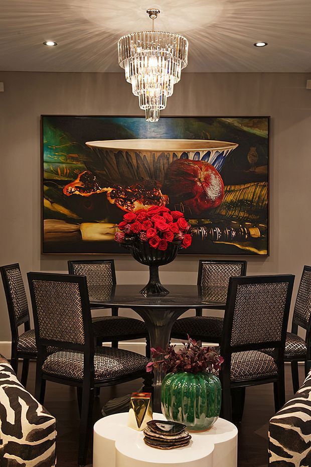 A richly styled dining area with a dark round dining table surrounded by patterned upholstered chairs. A large colourful still-life artwork anchors the wall, while a tiered crystal chandelier provides a dramatic focal point overhead. Black, cream and taupe finishes are accented by red floral arrangements, a green ceramic vase and decorative objects, with patterned lounge chairs visible in the foreground.