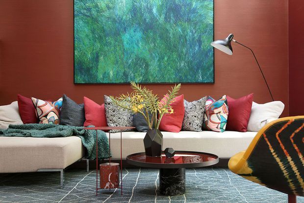 A richly layered living room with a large white sectional sofa arranged against a deep red feature wall. The space includes colourful patterned and solid cushions, a teal throw, a textured blue-grey rug, a low round coffee table with dark sculptural legs, a geometric vase arrangement, and a large abstract green-blue artwork. A slim black floor lamp and a partially visible patterned accent chair complete the eclectic, colourful scheme.