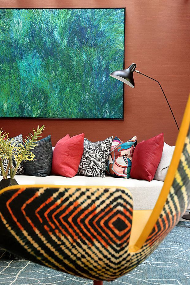 A richly layered living room vignette with a white sofa, colourful patterned cushions, a large abstract blue-green artwork, and a sculptural black wall lamp mounted on a warm reddish-brown timber wall. A prominent patterned lounge chair with a wooden frame occupies the foreground, set over a blue patterned rug. The styling is bold, eclectic, and highly colourful.