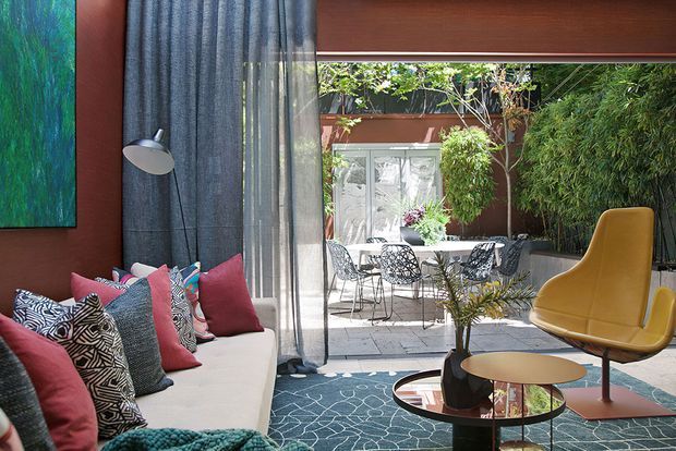 A richly styled contemporary living room opening onto a leafy paved courtyard. The room features a long cream sofa with colourful patterned cushions, a dark textured curtain, an abstract green artwork, a sculptural mustard lounge chair, round glass-and-metal coffee tables, and a patterned blue rug. Outdoor dining furniture and dense planting are visible beyond the large opening.