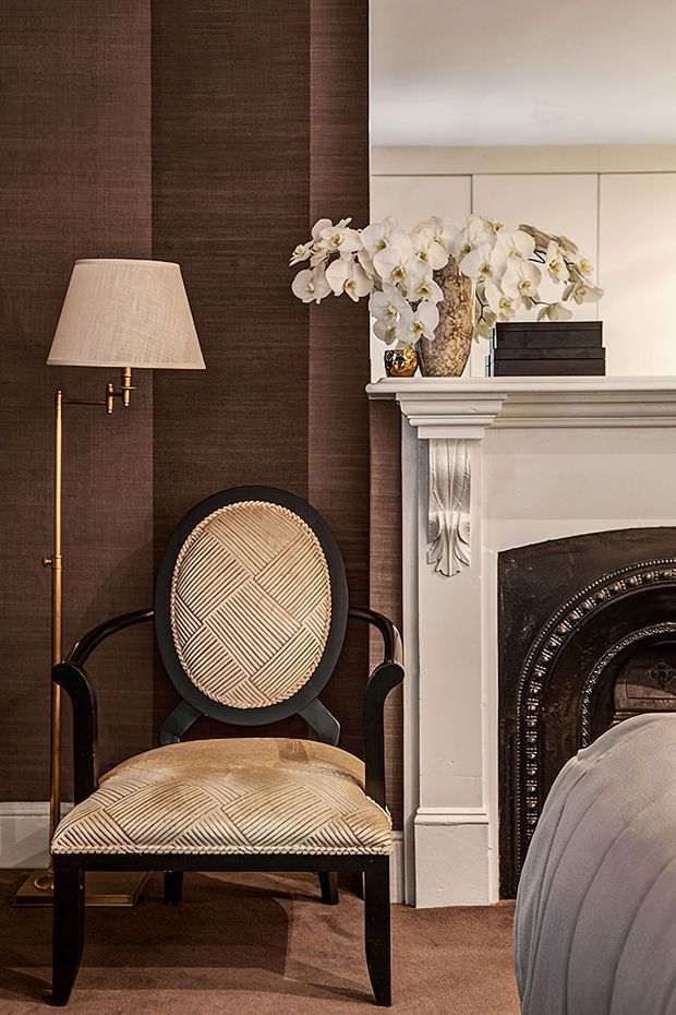 A refined bedroom corner featuring a black-framed upholstered armchair with woven geometric cream upholstery, a slim brass floor lamp, dark textured wall covering, and an ornate white fireplace mantel with floral arrangements and decorative objects. The edge of a bed is visible at right, confirming the bedroom setting. The composition has a layered, elegant, traditional feel with warm, muted lighting.