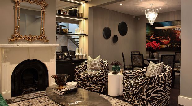 An eclectic, luxurious living room with a richly ornamented fireplace and ornate gold-framed mirror, dark built-in shelving, patterned zebra-print armchairs, a glossy stone coffee table, sculptural wall discs, and a crystal chandelier. The room has a warm, dramatic palette with layered decorative details and a dining area visible in the background.