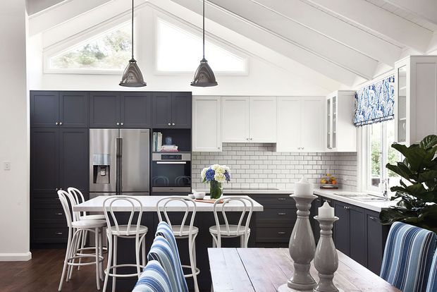 Bright open-plan kitchen and dining area with a vaulted white ceiling, exposed white beams, high clerestory windows, dark navy cabinetry, white subway-tile backsplash, stainless-steel appliances, a central island with bar seating, and a dining table with white metal chairs. Blue-and-white textiles and pendant lights add coastal character.