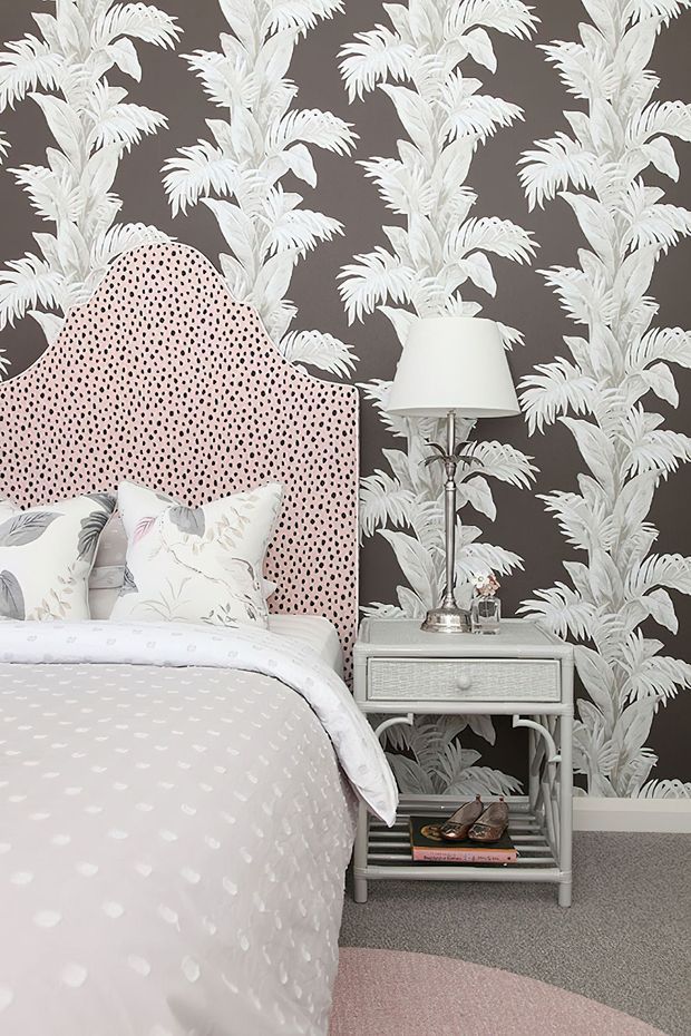 A styled bedroom corner featuring a bed with a tall scalloped pink upholstered headboard patterned with small dark spots, layered botanical-print pillows, and pale bedding. A white bedside table with open shelving holds a silver table lamp and books. The room is finished with dramatic charcoal wallpaper featuring repeating white tropical foliage, light carpet, and a round blush rug.