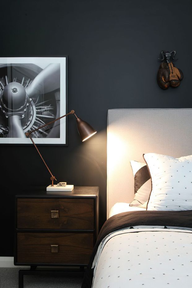 A moody bedroom vignette with a dark charcoal wall, upholstered light-grey headboard, white patterned bedding, and a dark timber bedside cabinet. A copper-toned articulated table lamp illuminates the bed, while monochrome framed artwork and a small decorative wall-mounted object add character.