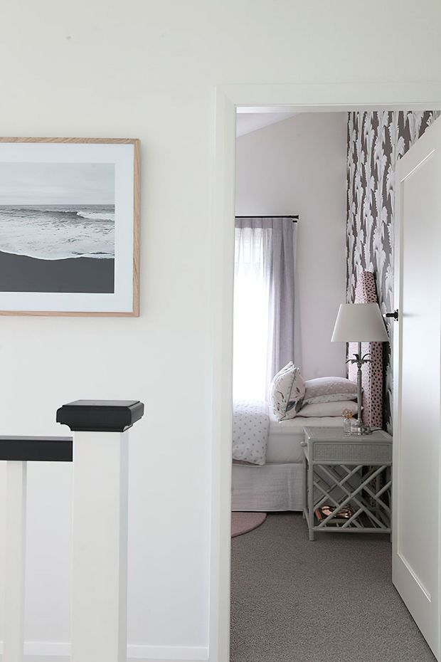A bright, white-painted hallway or landing looks through an open doorway into a softly styled bedroom. The bedroom contains a white upholstered bed with patterned bedding, sheer curtains, a grey lattice-front bedside table, a floor lamp, and a bold grey-and-white patterned feature wall. A framed monochrome ocean photograph and dark handrail accents appear in the foreground.