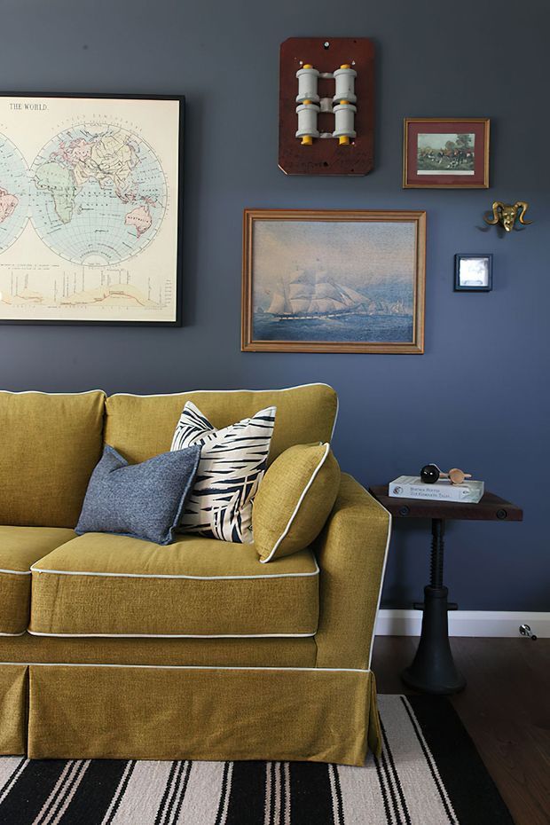 A richly styled living room vignette with a mustard-gold upholstered sofa, patterned and textured cushions, and a small dark pedestal side table. The deep navy-blue wall is arranged with framed artwork, a vintage world map, maritime paintings, a sculptural wall object, and a small framed picture. A black-and-cream striped rug sits over dark timber flooring, creating a warm, eclectic and slightly vintage atmosphere.