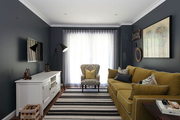 A compact, traditionally detailed living room with deep charcoal-blue walls, white decorative cornices, sheer curtains, a mustard-gold sectional sofa, patterned armchair, striped rug, and white built-in-style media cabinetry. Framed artwork, pendant-style floor lamps, and layered cushions add a refined, collected feel.
