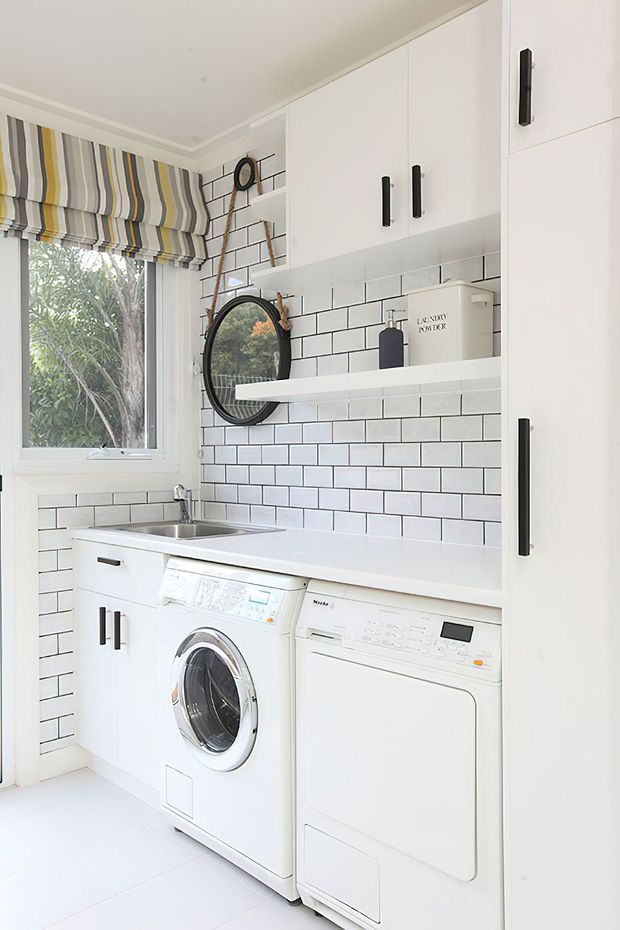 Bright, compact laundry room with white built-in cabinetry, a white benchtop, and white subway-tile walls with dark grout. Front-loading washing machine and dryer sit beneath the bench, with a stainless-steel sink beside them. A window with a striped Roman blind, black hardware, open shelving, and a round wall mirror add contrast and detail.