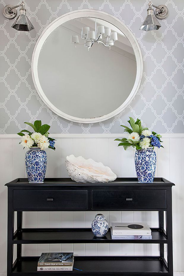 A styled interior vignette featuring a black open-shelved console with drawers, blue-and-white floral ceramic vases filled with flowers, and a large round white-framed mirror. The wall combines light grey geometric patterned wallpaper above white vertical wall panelling, with chrome wall sconces flanking the mirror. Decorative books and a sculptural shell sit on the console shelves.
