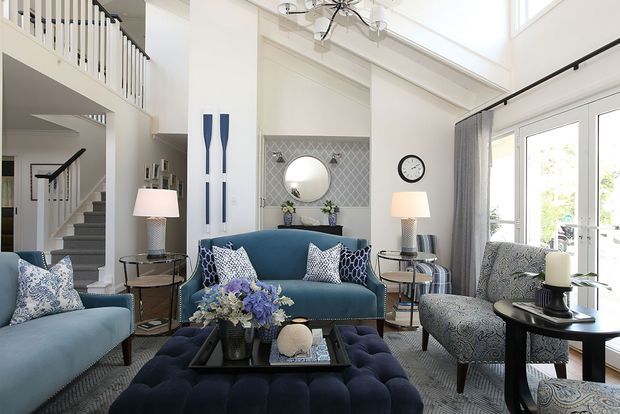 Bright, sophisticated living room with blue upholstered seating, a large tufted ottoman, patterned accent chairs and cushions, side tables, lamps, and a decorative fireplace or console wall with a round mirror. The room opens to a stairway and upper-level landing, with tall glazing and a high ceiling creating an airy, elegant interior.