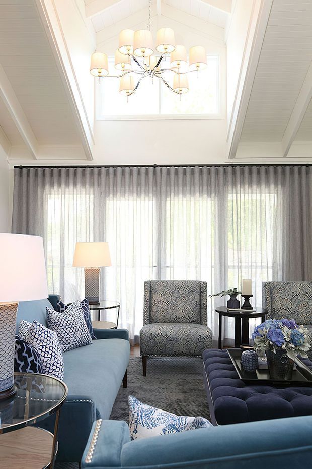 Bright, elegant living room with a vaulted white paneled ceiling, clerestory window, full-height sheer curtains, and a large decorative chandelier. The room is furnished with blue upholstered sofas, patterned armchairs and cushions, a tufted ottoman, glass-topped side tables, lamps, and floral styling. The palette is predominantly white, blue, grey, and silver with a refined traditional coastal feel.